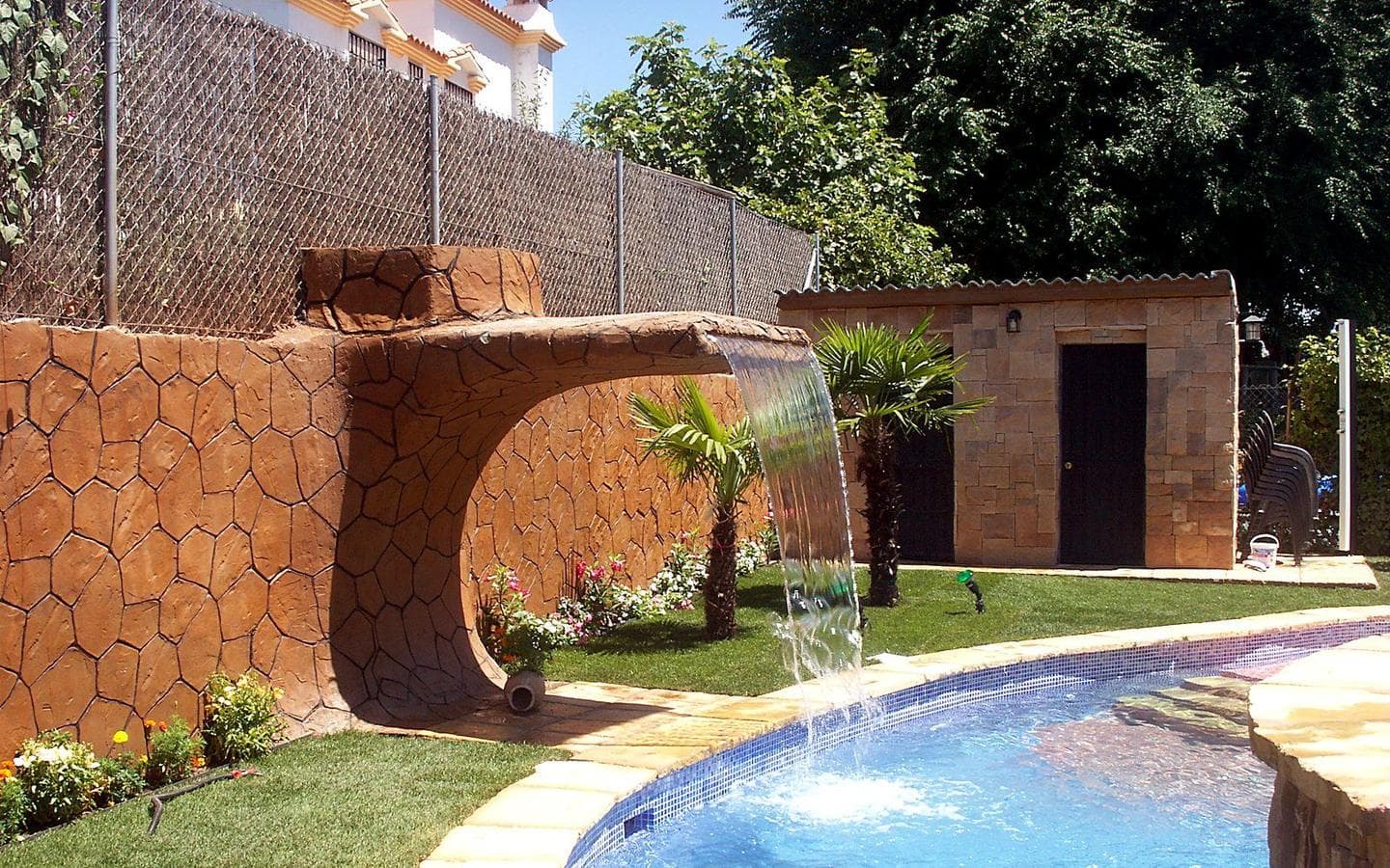 Muro de roca con una cascada en arco que cae sobre la piscina, entre palmeras, con la caseta de piedra al fondo