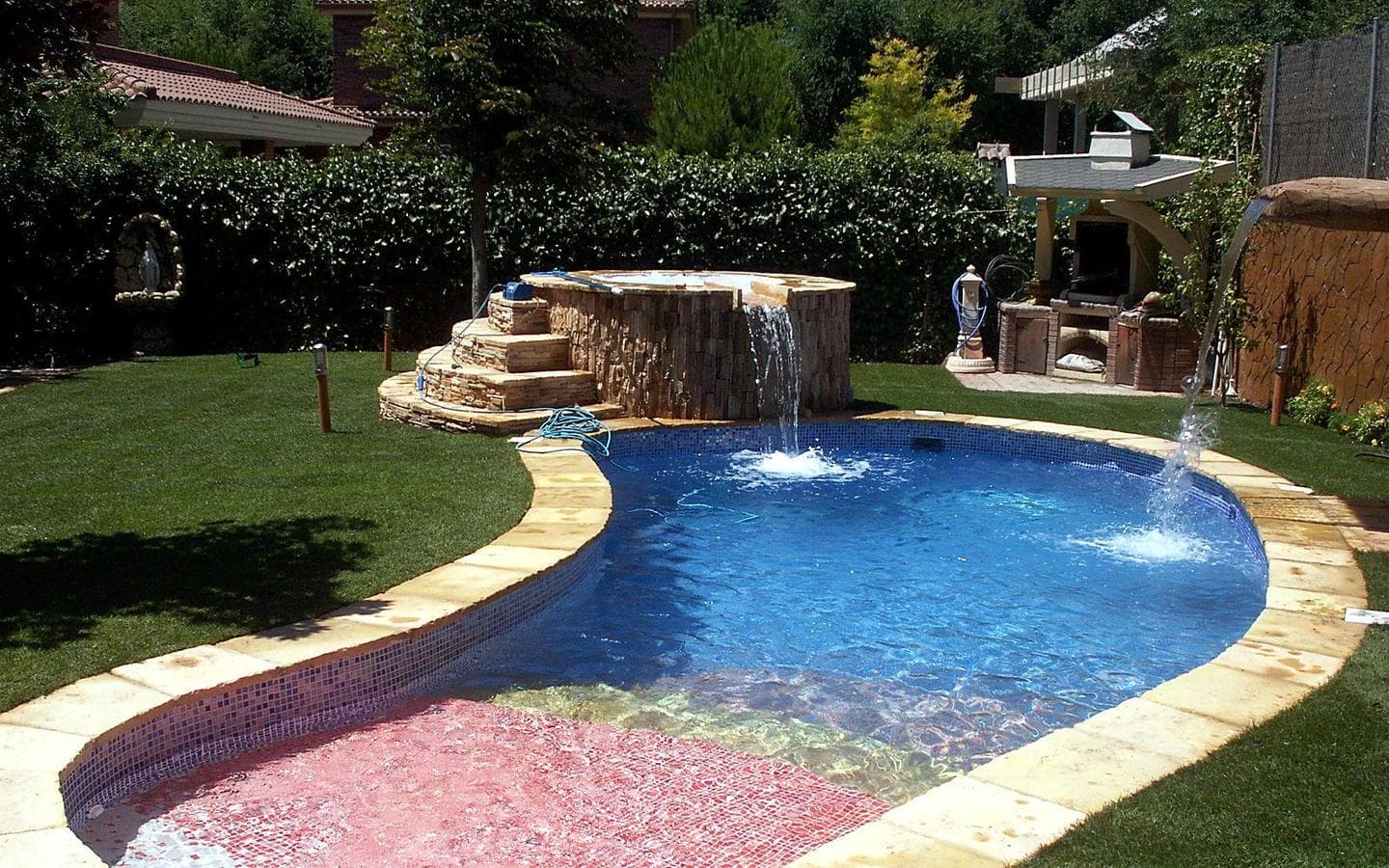 Piscina en forma de riñón con gresite azul y playa de gresite rosa; al fondo, spa elevado de piedra con cascada y chorro lateral, sobre césped