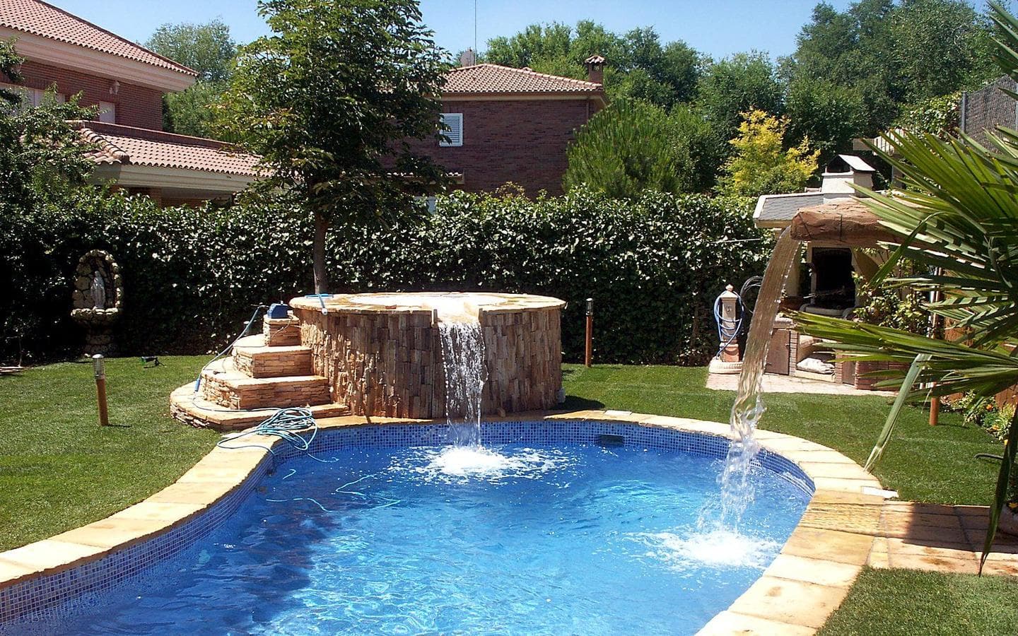 Piscina con el spa de piedra al fondo, su cascada y un chorro de agua, junto a una palmera