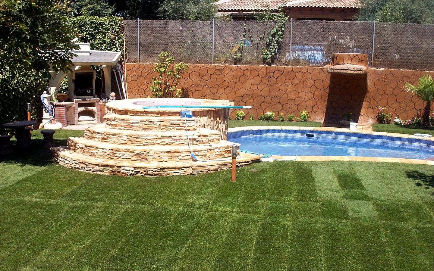 Vista general: spa circular elevado con escalones de piedra junto a la piscina, césped alrededor y muro de piedra al fondo
